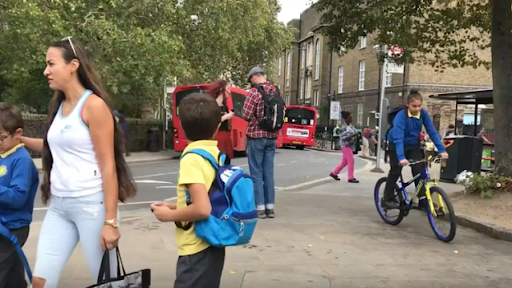Image of a street with a woman and man and four children, one on a bike. There are two red buses in the background.