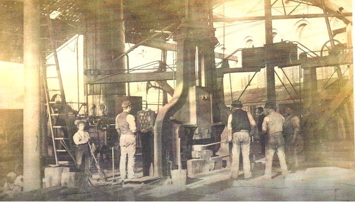 Sepia, yellow tinted image of hte inside of a factory with half a dozen men standing around.