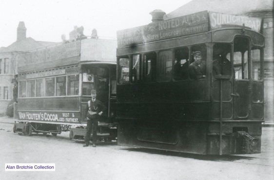 Monochrome image of a steam tram and trailer, with the driver looking out of the window and the conductor standng by the trailer.