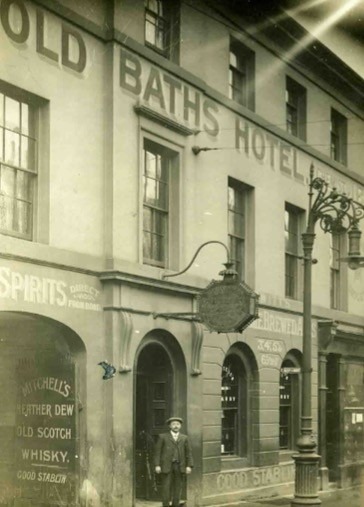 Old monochrome image showing a man standing infront od rhw Ols Baths Hotel