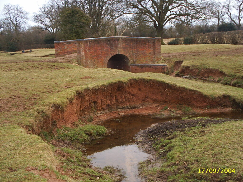 Colour photograph of a small winding river and a bridge.