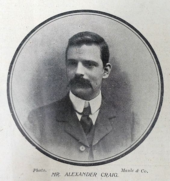 Early 1900s black-and-white portrait of a man with short dark hair and a thick mustache, wearing a high-collared shirt, tie, and dark three-piece suit, framed in a circular border.
