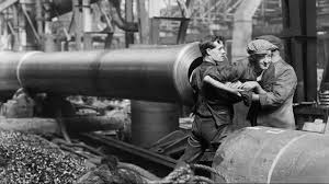A monochrome image showing a woman being pulled out of a large naval gun barrel with two men helping to remove her. The woman is wearing a flat cap