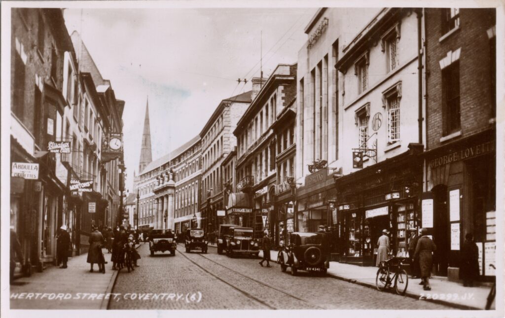 Old black and white photo of a busy street with old cars lining the right hand side, a parked bickcly and a spire in the distance.