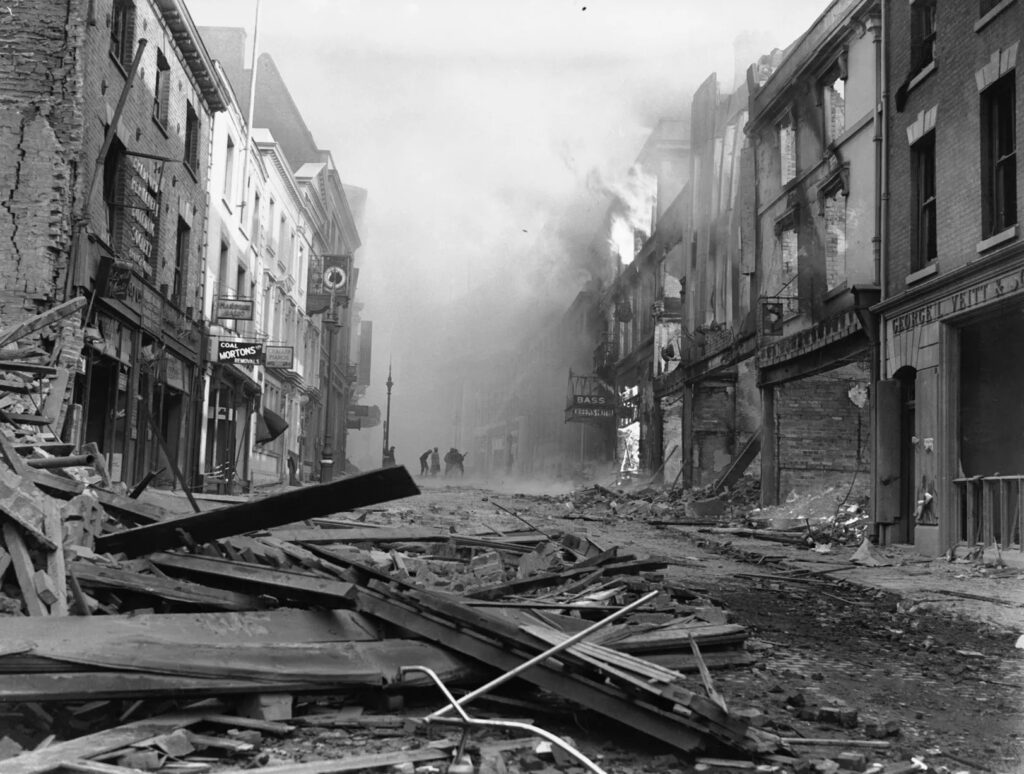Black and white photo showing the destruction of the street after the bombing. there is damage in the foreground, and wrecked building to the right and smoke behind.