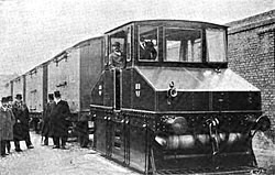 Historic black-and-white photo of an early petrol-powered locomotive pulling freight wagons, with several men in period clothing standing nearby.