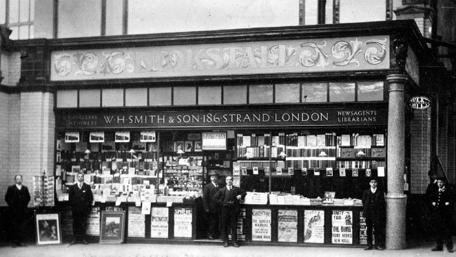 Black and white photo of a busy shop front with the sign "WH Smith and son 186 Strand London with two men in the middle and other men at the ends.