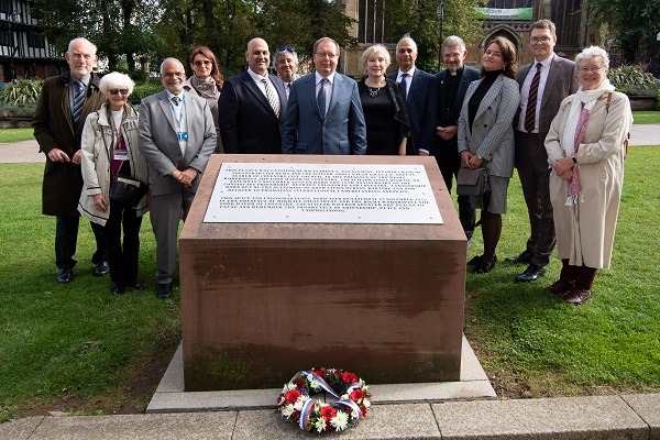 Phiotograph of a red sandstone plinth with a plaque on the top. Thirteen people are standing arounnd the monument and a wreath is front of it.