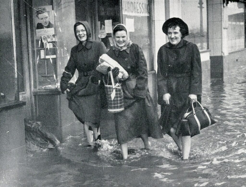 Monochrome image showing three women in raincoats wading through flood water.