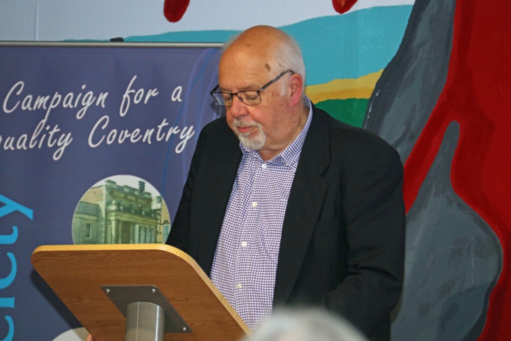 Phoitograph of Peter Walters reading from a lecern with a Coventry Society poster behind.