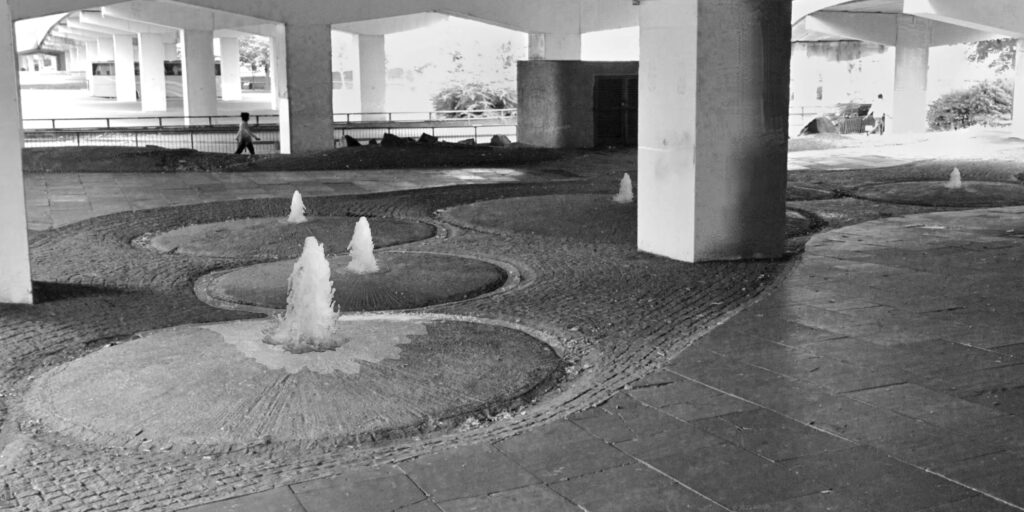 Monochrome photograph showing five crater like structures gushing water in the area under the Ring Road. The columns of the ring road run into the distance.