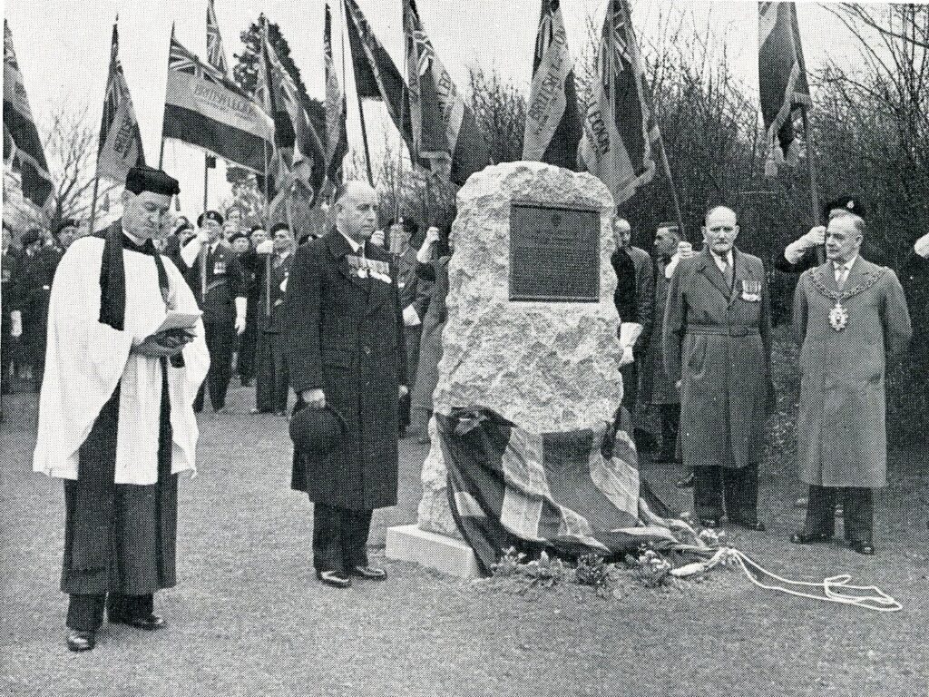 Monochrome photograph showing a stone memorial with plaque, sourrounding by civic dignatories, church personell and soldiers with flags.