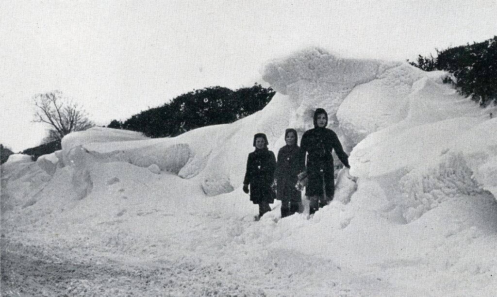 Monochrome image showing three children standing in deep snow.